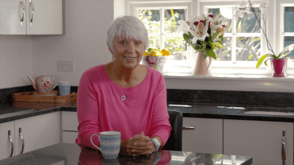 A happy careline alarm user wearing a Telecare24 fall alarm and sitting in her kitchen with a cup of tea